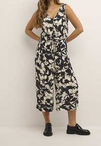 Jumpsuit floral con un patrón en negro y crema, escote en V, cintura ceñida y piernas anchas y recortadas. Modelo luciendo zapatos de plataforma negros.