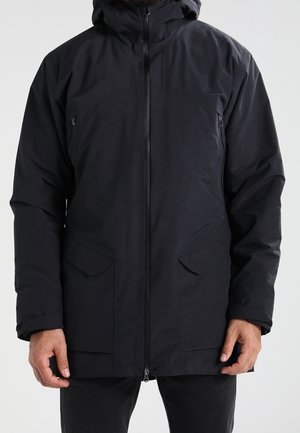 Mand iført en sort hættetrøje vinterjakke med frontlynlås og flere lynlåse- og klaplommer, stående mod en ensfarvet baggrund.