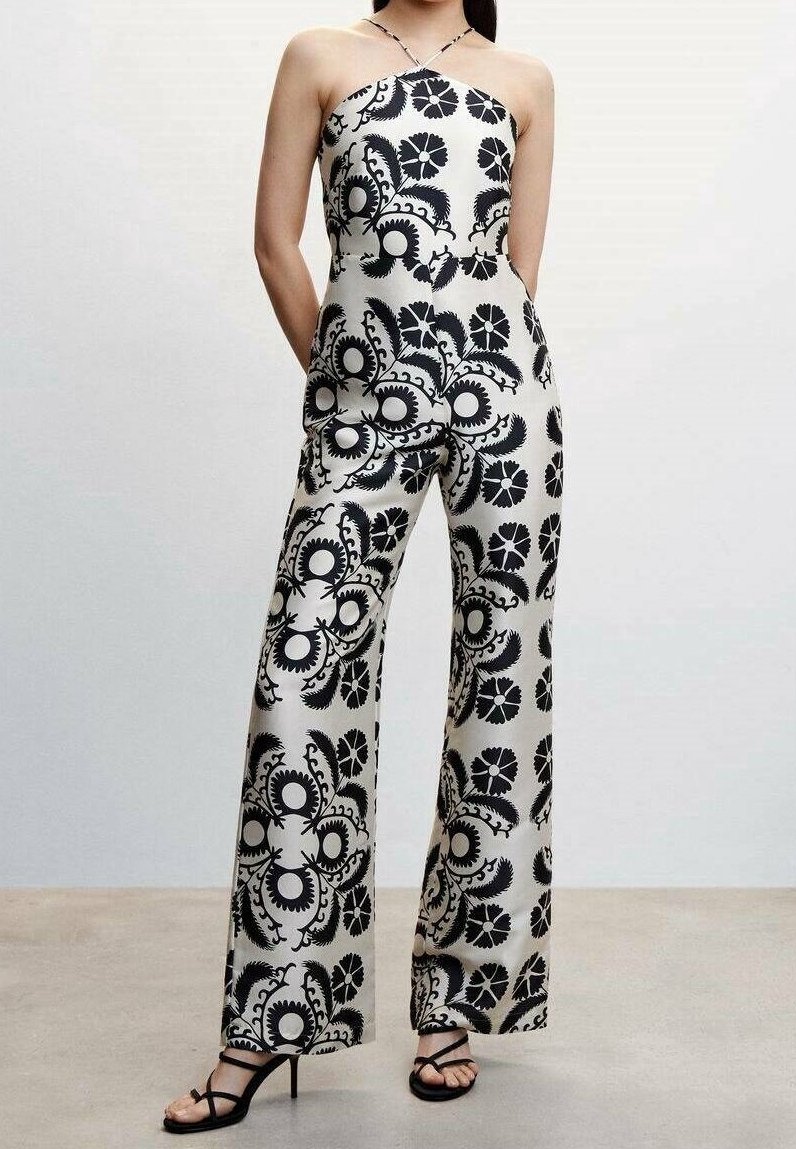Femme portant une combinaison blanche à col licou avec un motif floral noir et des sandales à talons hauts noires à lanières, debout les mains dans les poches.