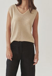 Beige stickad ärmlös topp med V-ringning, med en strukturerad yta. Kombinerad med mörka tygbyxor som har en avslappnad passform.