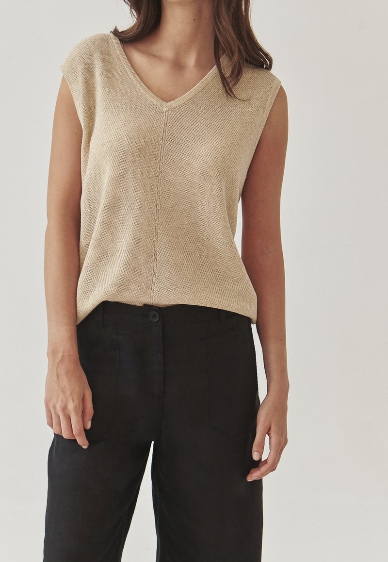 Beige stickad ärmlös topp med V-ringning, med en strukturerad yta. Kombinerad med mörka tygbyxor som har en avslappnad passform.