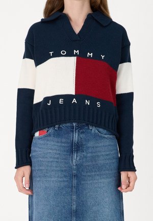 Kobieta w swetrze w blokowe kolory: granatowy, biały i czerwony z napisem "Tommy Jeans", w połączeniu z niebieską dżinsową spódnicą.