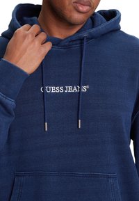 Felpa con cappuccio blu navy realizzata in tessuto morbido, con tasca frontale, stringhe con punte in metallo e "GUESS JEANS" ricamato in bianco.