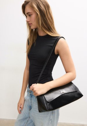 Mladi žena s dugom plavom kosom nosi crnu majicu bez rukava i svijetloplave traperice, držeći crnu kožnu torbu preko ramena.