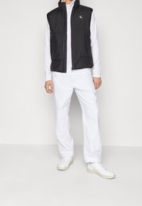 Schwarze ärmellose Weste mit einem Frontreißverschluss und einem Logo auf der Brust, kombiniert mit weißem Langarmshirt und weißen Hosen, dazu getragen mit hellen Schuhen.