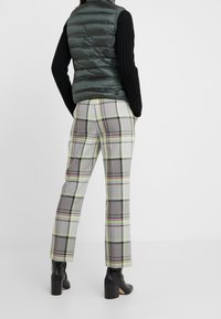 Grüne Steppweste mit hohem Kragen über einem schwarzen Strickpullover. Graue Karohosen mit rosa und grünen Linien, kombiniert mit schwarzen Absatzstiefeln.