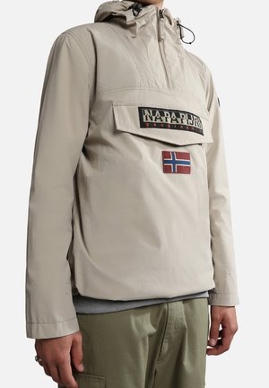 Beige Kapuzenjacke mit Frontklapptasche mit der Aufschrift "Napapijri Geographic" und einem norwegischen Flaggen-Patch über olivgrüner Hose.