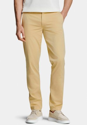 Persoon draagt beige chino's en lichtbeige veterschoenen, staat met één hand in de zak, bij de taille bijgesneden.
