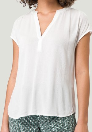 Blouse blanche à manches courtes avec décolleté en V, en tissu texturé, et coupe décontractée, associée à un pantalon vert à motifs.