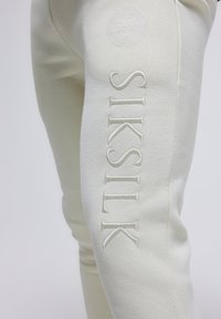 Beiga joggers med en mjuk textur, med en broderad "SIKSILK"-logotyp på sidan och ett lejon-emblem ovanför.