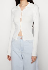Top bianco a maniche lunghe con righe e colletto, chiusura centrale con bottoni e apertura sul ventre; abbinato a jeans a vita alta di colore blu chiaro.