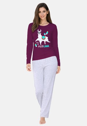 Donna che indossa una maglietta a maniche lunghe viola con grafica bianca di lama e testo "not my problama", abbinata a pantaloni grigio chiaro, in piedi scalza.