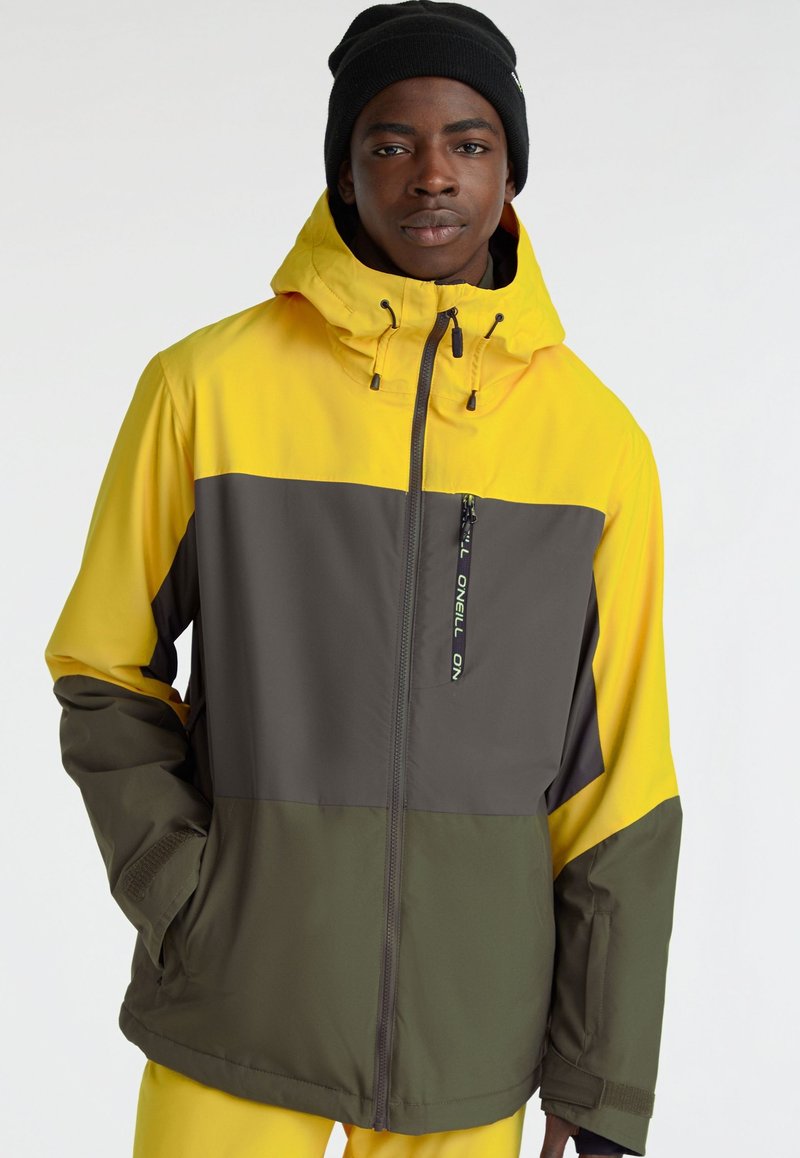 Mężczyzna w kurtce w kolorze blokowym w odcieniach żółtego, szarego i oliwkowego z kapturem oraz czarnej czapce na jednolitym tle.