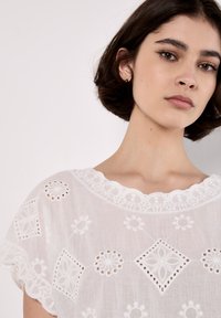 White embroidered blouse featuring scalloped neckline, floral patterns, and cut-out details. Made from lightweight fabric with a textured appearance.