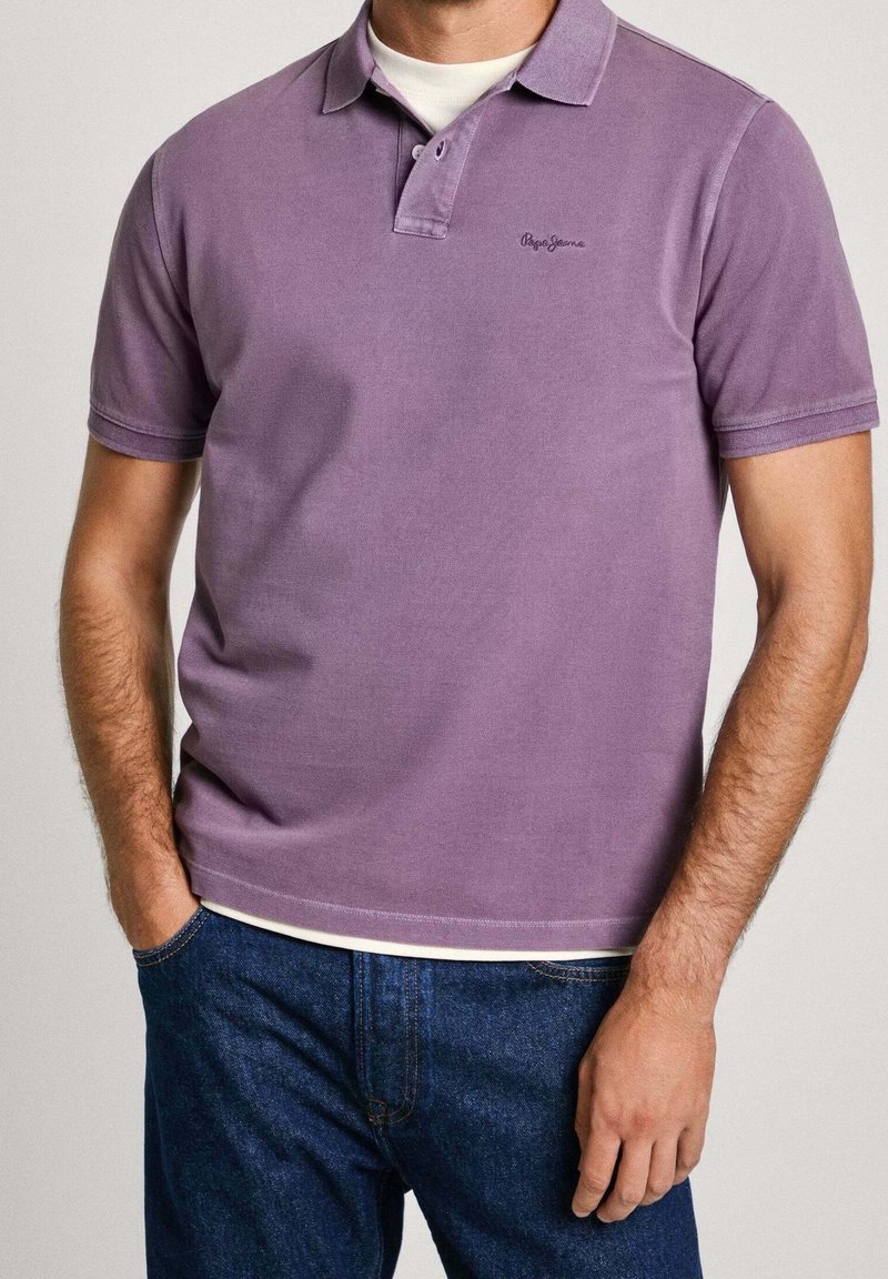 Homme portant un polo à manches courtes lavande par-dessus un sous-vêtement blanc avec un jean bleu, les mains dans les poches, visible du cou aux hanches.