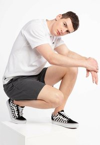 Zwart-witte geblokte sneakers met witte zolen, gecombineerd met grijze shorts en een wit T-shirt. Model poseert in een hurkende positie.