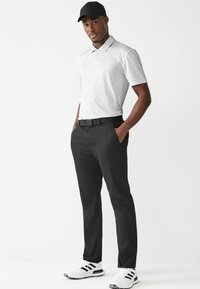 Texturerad grå pikétröja, svarta byxor och vita sportskor med svarta detaljer. Modellen bär en svart keps; armarna avslappnade, händerna i fickorna.