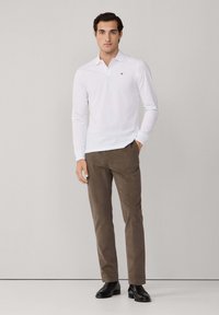 Mann steht in einem weißen Langarm-Polohemd, braunen Hosen und schwarzen Geschäftsschuhen vor einem schlichten hellen Hintergrund.
