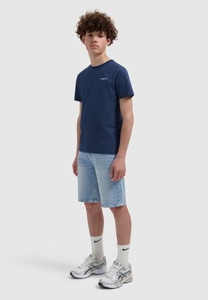 Tiener met krullend haar, gekleed in een marineblauw T-shirt, lichte spijkershorts, witte Nike-sokken en zilveren hardloopschoenen, staand tegen een effen achtergrond.