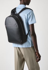 Zaino nero con un pannello inferiore testurizzato, design elegante, zip argentata, indossato con una camicia azzurra chiara. Dotato di spallacci imbottiti e regolabili.