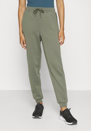 Personne portant un pantalon de survêtement vert olive à cordon et des chaussures de sport noires, debout sur un fond de sol et mur blanc uni.