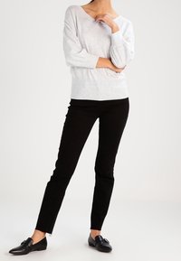 Grauer Pullover mit V-Ausschnitt und weiten Ärmeln, kombiniert mit schwarzen, schmal geschnittenen Hosen und schwarzen Slippern, die ein einfaches Design aufweisen.