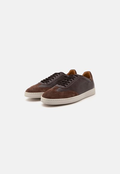 Paire de baskets basses pour homme en cuir marron foncé et suède avec des lacets noirs et des semelles en caoutchouc blanches sur un fond uni.