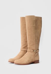 Polven korkeudella olevat beigenväriset mokkanahkasaappaat, joissa on pyöreä kärki, kullanvärisillä metalliosilla koristeltu remmi ja matala puukoroke.