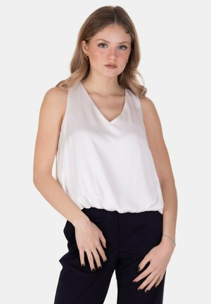 Giovane donna con capelli biondi che indossa una camicetta bianca senza maniche e pantaloni neri, in piedi davanti a uno sfondo chiaro e uniforme.