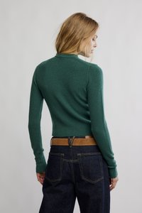 Grønn ribbestrikket genser med lange ermer, tettsittende design og høy hals. Kombinert med mørke denimjeans og et brunt belte. Ensfarget bakgrunn.