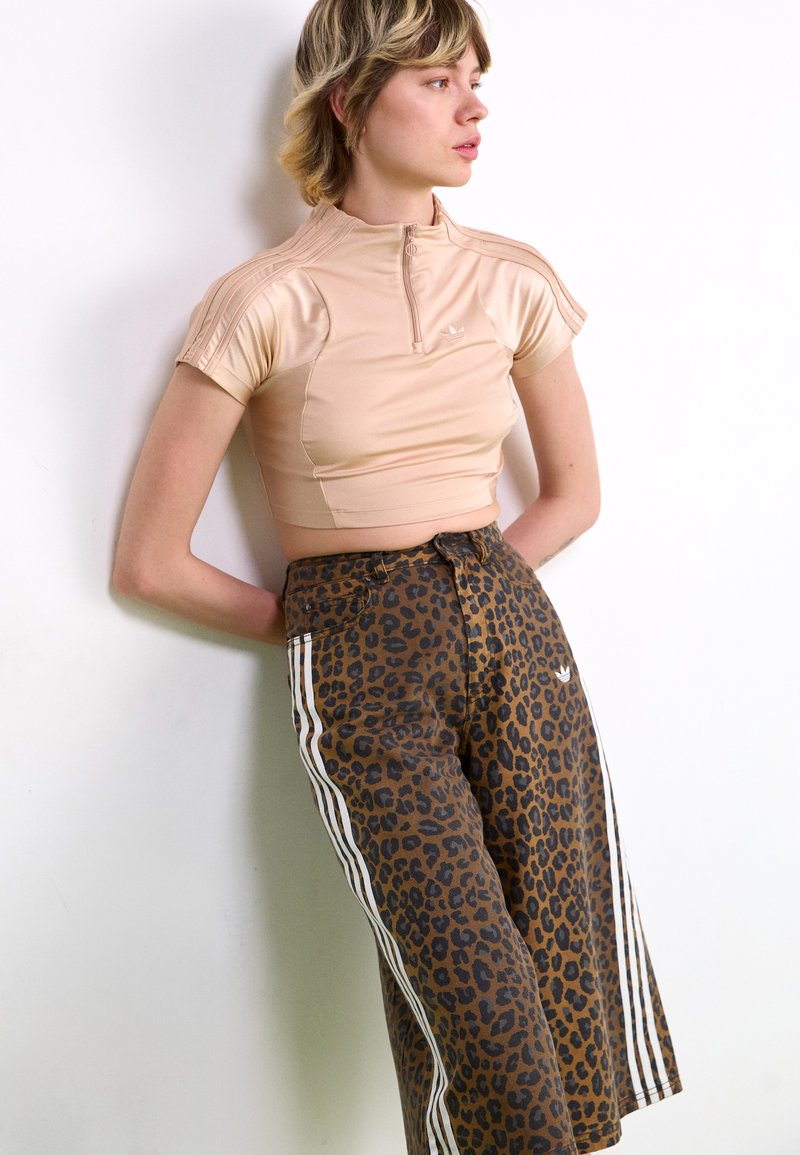 Young person with short hair wearing beige cropped zip-up top and brown leopard print pants with white stripes, leaning against a white wall.
