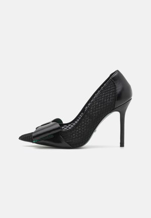 Black high-heeled shoe with a pointed toe, featuring a smooth leather front and textured mesh sides, adorned with a large bow detail.