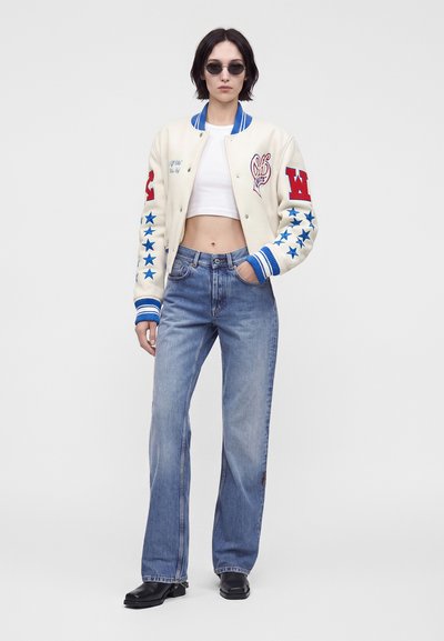 Γυναίκα με στρογγυλά γυαλιά ηλίου, λευκό cropped varsity μπουφάν με μπλε αστέρια και κόκκινες μπαλώματα, μπλε τζιν ψηλής μέσης και μαύρες μπότες.