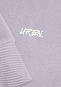 Sudadera de color lila con puños de canalé, que presenta el texto "URBN." en blanco impreso en estilo pincel. Textura suave, ajuste casual, sin patrones adicionales.