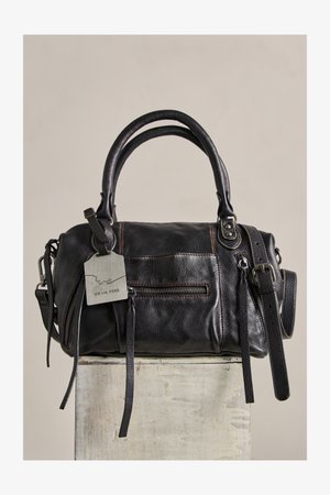 Černá kožená kabelka s dvojitými držadly, přední kapsou na zip, připevněnou visačkou a nastavitelným ramenním popruhem, položená na světlém bloku.