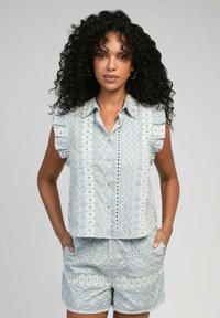 Chemise bleu clair à manches courtes avec des épaules froncées. Présente des broderies blanches, des motifs géométriques, et un short assorti avec des poches.