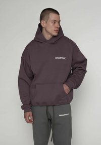 Bordowa bluza z kapturem z kieszenią kangurka i białym napisem "MULTIPLY" na piersi. Miękki, gruby materiał, zestawiona z szarymi spodenkami dresowymi.