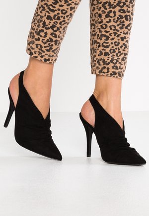 Fødder iført sorte spidse slingback-højhælede sko kombineret med beige leopardprint-korte bukser på en hvid gulv.