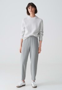 Grauer gerippter Pullover mit hohem Kragen, kombiniert mit hellgrauen, schmal zulaufenden Hosen. Weiße Sneakers mit grauen Akzenten runden das Outfit ab.