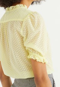Blusa gialla traslucida con piccoli motivi floreali bianchi, maniche a sbuffo e polsini frillati, caratterizzata da un colletto arricciato e tessuto testurizzato.