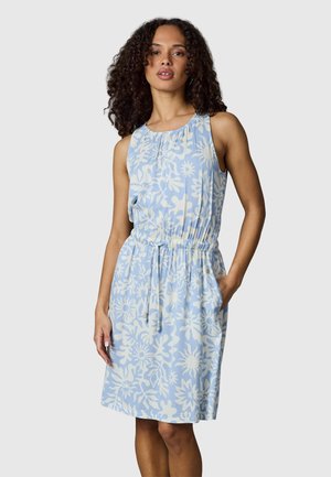 Vrouw met krullend haar die een lichtblauwe mouwloze jurk met wit bloemenpatroon en elastische taille draagt, staand met één hand in de zak.