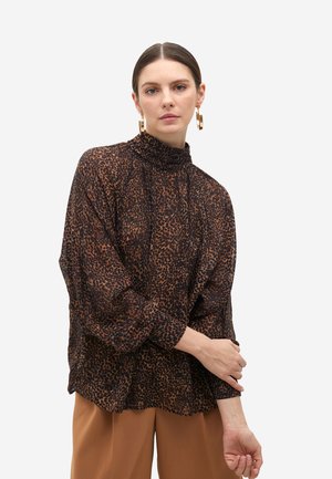Blusa de estampado de leopardo negro y marrón con cuello alto y mangas vaporosas, hecha de una tela ligera. Combinada con pantalones color arena.