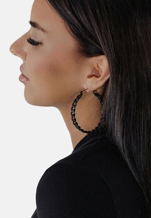 Frau mit langen dunklen Haaren, die großen schwarzen geflochtenen Creolenohrring trägt, nach links blickend in schwarzer Kleidung vor weißem Hintergrund.