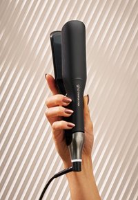 Main tenant un fer à lisser ghd chroma max noir avec des ongles couleur or rose sur un fond beige texturé.