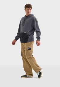 Felpa con cappuccio grigia e nera con tasca frontale, abbinata a pantaloni cargo color khaki dotati di più tasche e un patch con logo. Sneakers con dettagli neri, bianchi e verdi.