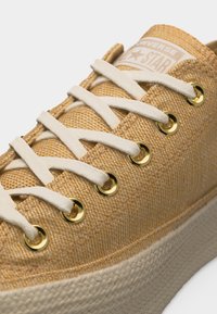 Καφέ πλεκτό αθλητικό παπούτσι Converse με λευκά κορδόνια, χρυσό μάτι και παχύ, υφή, μπεζ ελαστικό πέλμα.