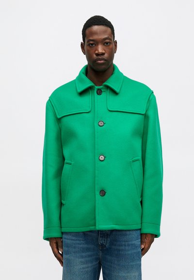 Chaqueta de mezcla de lana verde con cuello estructurado, cierre frontal de botones y dos bolsillos laterales, combinada con vaqueros de mezclilla azules.