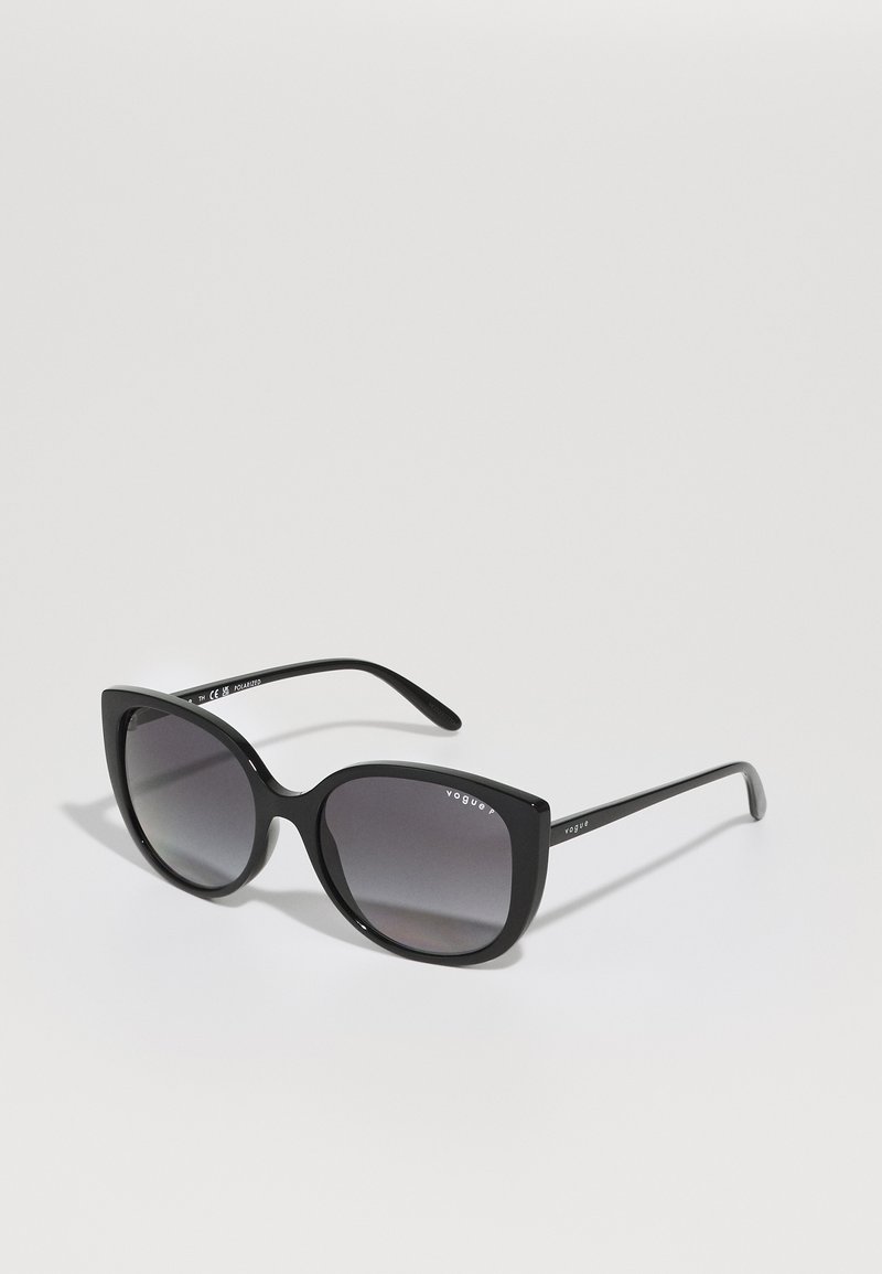 Óculos de sol oversized pretos com formato de olhos de gato, com lentes cinza degradê e a marca "Vogue" nas hastes. Armação de plástico suave.