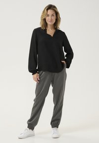 Black long-sleeve top with a V-neck, paired with gray tapered trousers and white sneakers. The fabric has a textured finish and gathered cuffs.