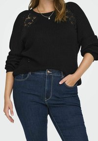 Svart stickad tröja med spetsdetaljer på axlarna, i kombination med mörkblå jeans. Tröjan har uppvikta ärmar och en avslappnad passform.